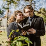 Man in Black Suit Jacket Reading a Bible Beside a Grieving Woman in Black Coat