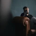 A man is sitting on a sofa with his hands on his head