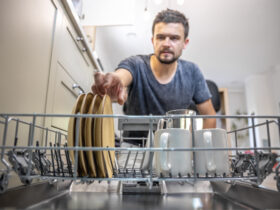 A man in front of an open dishwasher takes out or puts down dishes