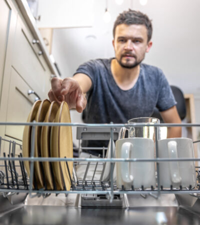 A man in front of an open dishwasher takes out or puts down dishes