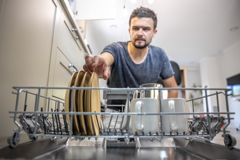 A man in front of an open dishwasher takes out or puts down dishes