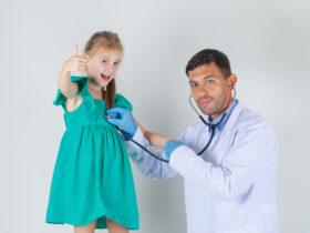 Male doctor in white coat listening heartbeat while child showing thumb up