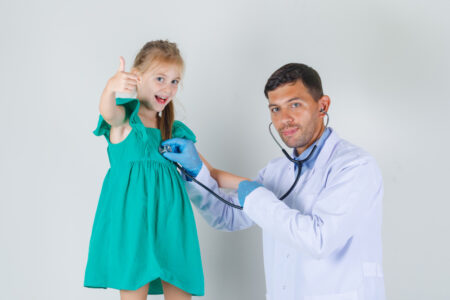 Male doctor in white coat listening heartbeat while child showing thumb up